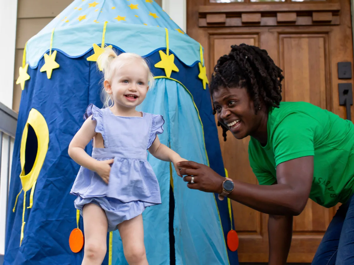 Safety tips for sitters: A smiling SitterTree sitter wearing a green SitterTree T-shirt playing with a happy toddler near a colorful play tent on a porch, capturing a fun and caring moment