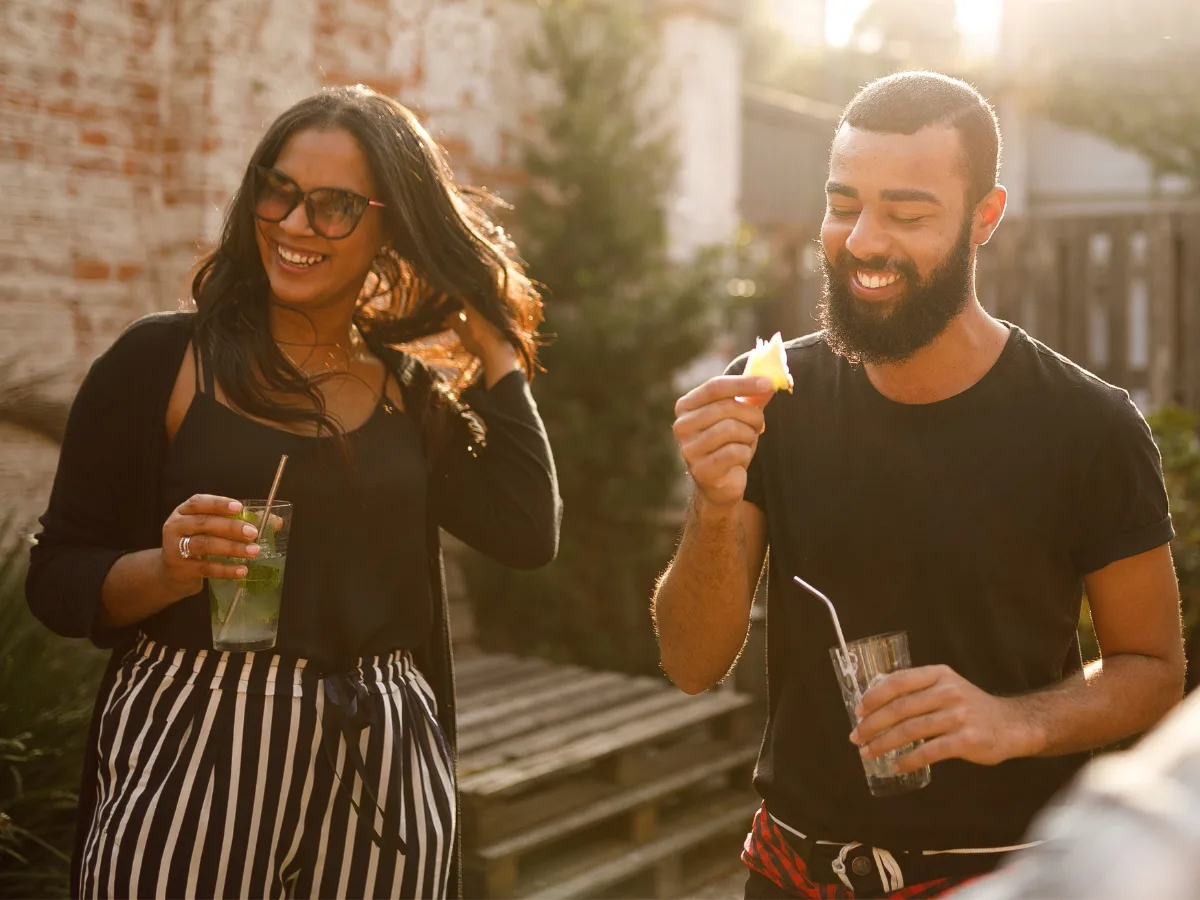 date night ideas in Charlotte: A smiling couple enjoying a casual outdoor date. The woman holds a cocktail while adjusting her hair, and the man laughs while holding a drink in one hand and his garnish in the other.