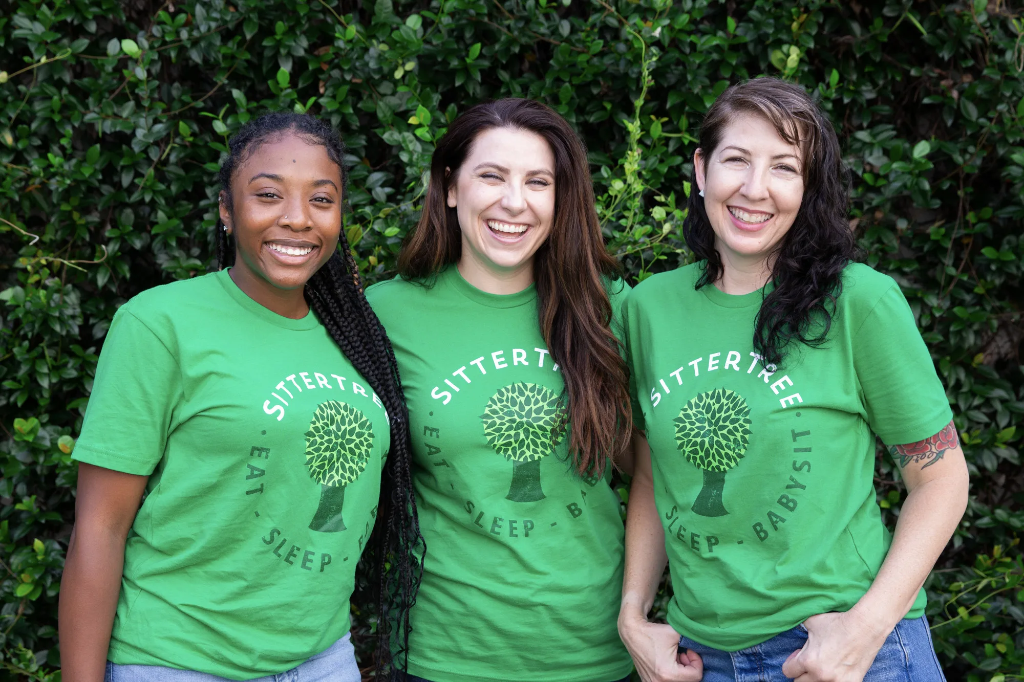 How much to pay a babysitter per hour: Three smiling women wearing green SitterTree t-shirts standing together in front of a lush green leafy background, radiating warmth and camaraderie